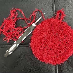May include: A red crocheted round dishcloth with a black crochet hook and red yarn.