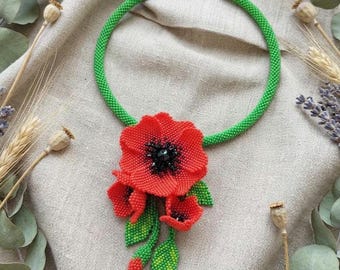 May include: A beaded necklace featuring a vibrant red poppy pendant with black accents and green leaves. The necklace has a bright green beaded collar. The poppy pendant is the focal point, showcasing intricate beadwork. A unique floral accessory.