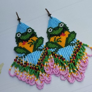 May include: A pair of beaded frog earrings with a blue, green, pink, white, and yellow color scheme. The earrings feature a fringe design and are made with small beads.