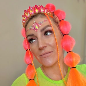 May include: A woman with bright orange hair styled in bubble braids and long strands. She wears a jeweled crown and face gems. The crown and face gems are pink and iridescent. She is wearing a neon green top.