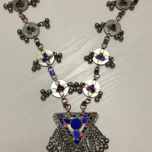 May include: A silver necklace with a triangular pendant featuring colorful enamel and a fringe of silver beads. The necklace is made up of circular silver pieces with colorful enamel accents and silver beads.
