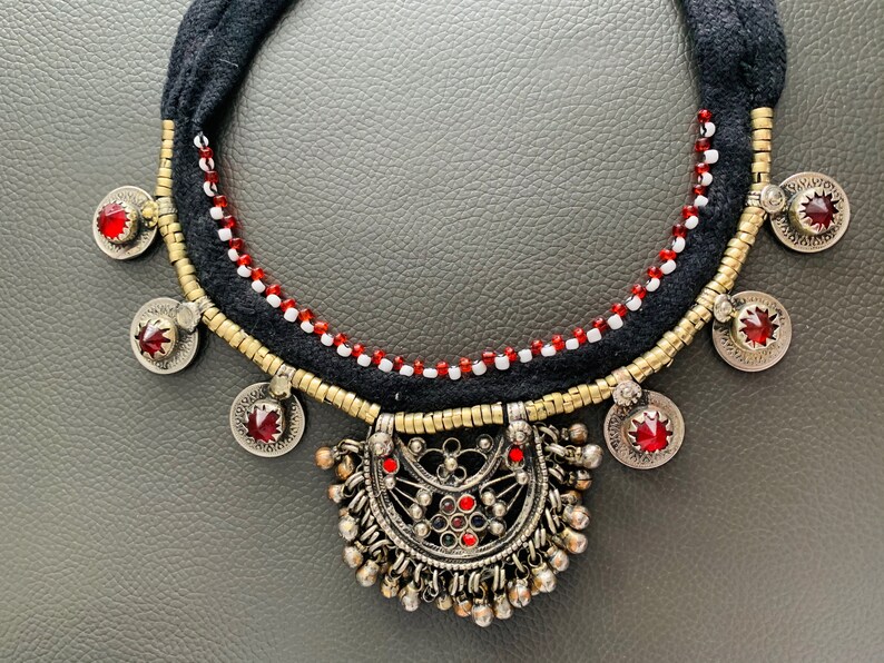 May include: A black and gold necklace with red and white beads. The necklace has a large, ornate pendant with a red stone in the center.