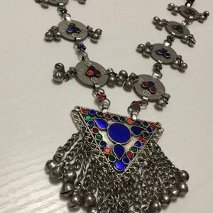 May include: A silver necklace with a triangular pendant featuring blue, red, and green enamel. The pendant is adorned with small bells and hangs from a chain with circular charms.