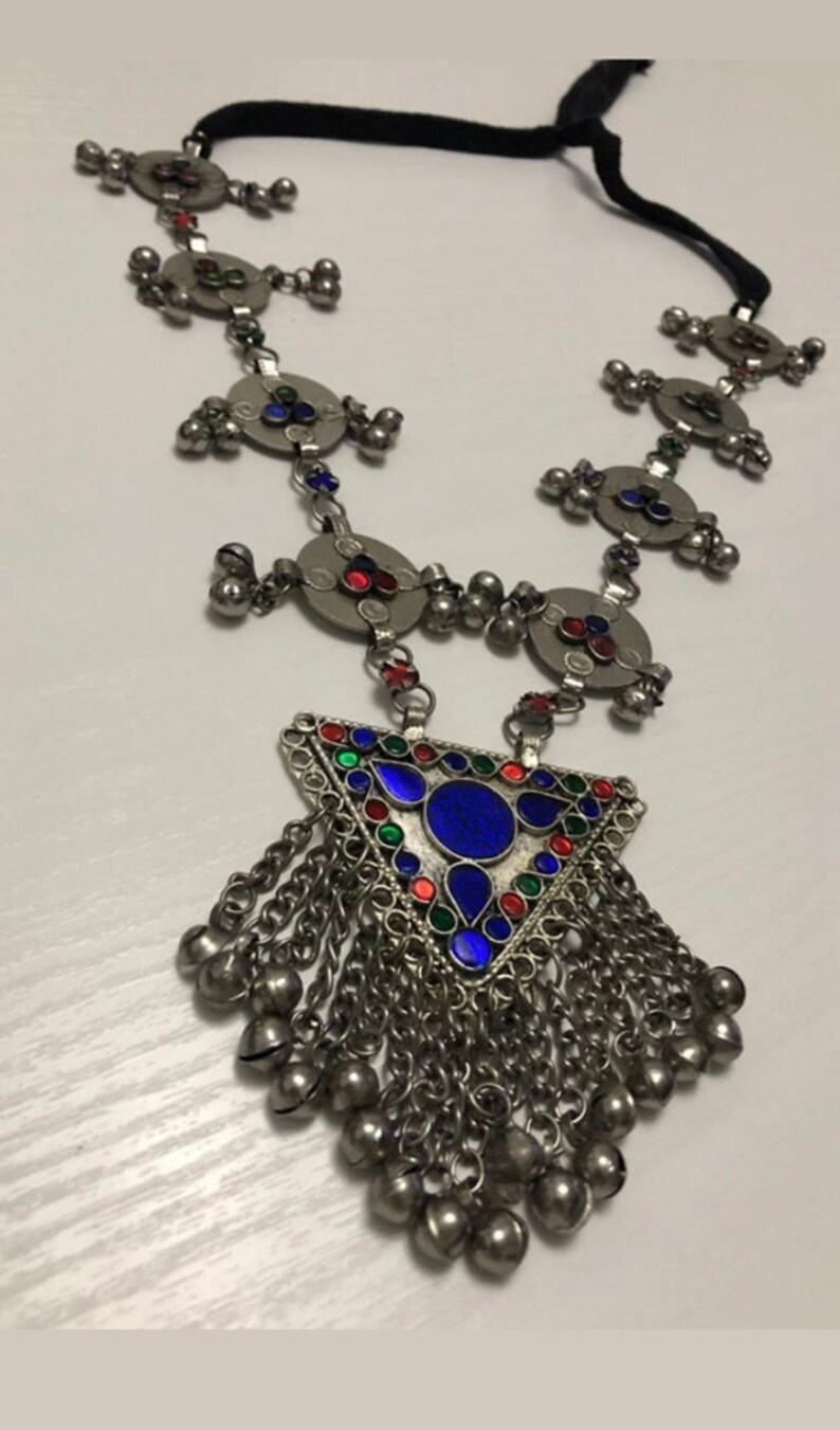 May include: A silver necklace with a triangular pendant featuring blue, red, and green enamel. The pendant is adorned with small bells and hangs from a chain with circular charms.