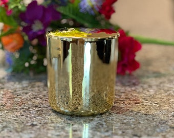 Puede incluir: Un recipiente cilíndrico para velas con un acabado metálico dorado reflectante. La parte superior del recipiente cuenta con una tapa lisa y plana que refleja los colores vibrantes de un ramo de flores amarillas, moradas, rojas y naranjas.