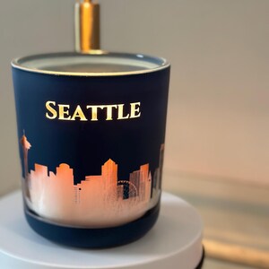 Puede incluir: Una vela azul con una silueta dorada del horizonte de Seattle y la palabra "SEATTLE" en letras doradas. La vela está sobre una base circular blanca.
