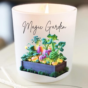 Könnte beinhalten: Eine brennende Kerze in einem Milchglas mit dem Schriftzug "Magic Garden". Das Glas zeigt eine Miniaturgartenszene mit bunten Gemüsesorten. Zwei Streichhölzer mit blauen Spitzen befinden sich im Vordergrund.