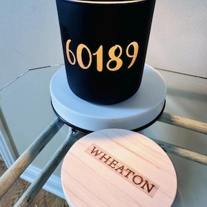 May include: A black candle holder with gold lettering reading "60189" sits on a white base. A wooden lid with the word "WHEATON" is below. The candle holder is on a glass surface.