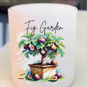 Puede incluir: Un portavelas cilíndrico blanco con una ilustración en acuarela de una higuera en una maceta de madera. El árbol está cargado de higos maduros. Las palabras "Fig Garden" están escritas en una elegante escritura sobre el árbol.