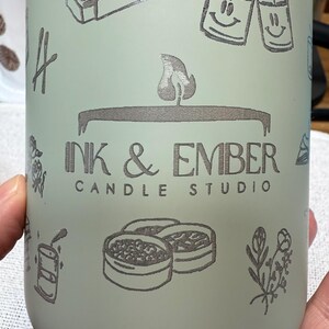 May include: A matte sage green cylindrical container featuring dark gray line art illustrations of matchboxes, smiling candles, and floral sprigs. The center displays the text INK & EMBER CANDLE STUDIO with a flame icon above the lettering.