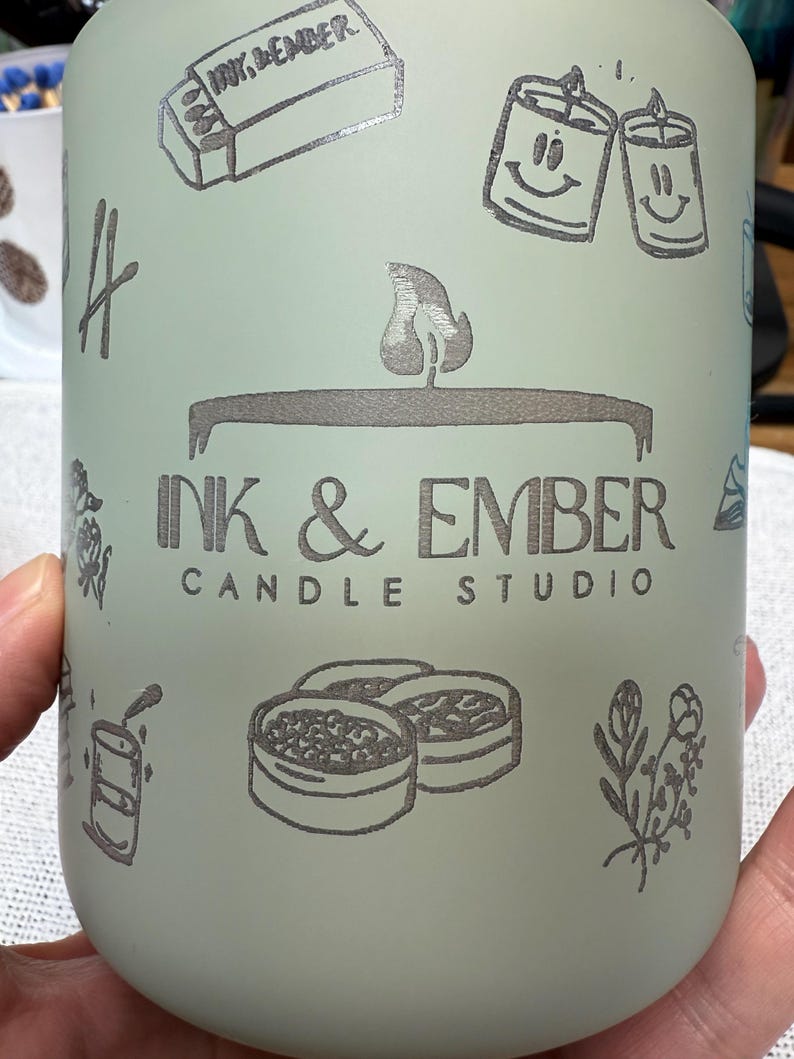 May include: A matte sage green cylindrical container featuring dark gray line art illustrations of matchboxes, smiling candles, and floral sprigs. The center displays the text INK & EMBER CANDLE STUDIO with a flame icon above the lettering.