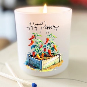 May include: A white candle with the text "Hot Peppers" and a colourful illustration of chilli peppers growing in a wooden planter. Two matches with blue tips are in the foreground. The candle is lit.