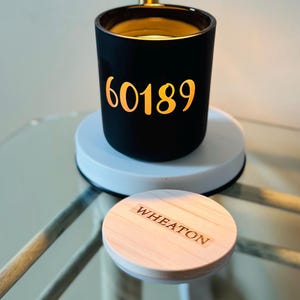 May include: A black candle holder with the number 60189 in gold lettering. The candle holder sits on a white base, with a wooden lid that reads "WHEATON". The candle holder is on a glass table.