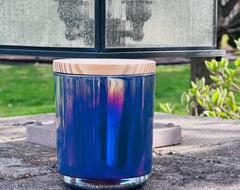 May include: A blue candle jar with a wooden lid and a silver base. The jar has a glossy, iridescent finish. The candle jar is on a stone surface. The jar's height is approximately 10 cm.