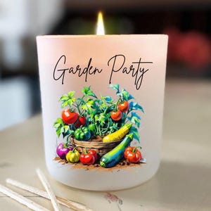 Könnte beinhalten: Eine brennende Kerze in einem Glas mit Milchglas-Effekt und dem Schriftzug "Garden Party". Das Glas zeigt eine farbenfrohe Illustration eines Korbes voller Tomaten, Zucchini und anderem Gemüse. Streichhölzer mit blauen Köpfen sind im Vordergrund.