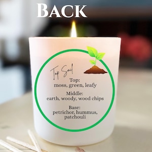 Puede incluir: Una vela blanca encendida con el texto "Top Soil" y un diseño circular verde. La etiqueta de la vela enumera las notas de la fragancia: musgo, verde, frondoso, tierra, amaderado, virutas de madera, petricor, hummus y pachulí.