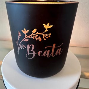 May include: Black candle jar with a floral design and the name "Beata" in gold lettering.