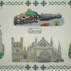 May include: A cross-stitch embroidery depicting various landmarks of Devon, England. The design features a lighthouse, a church, a town, and a clock tower. The word "Devon" is stitched in the centre of the design.