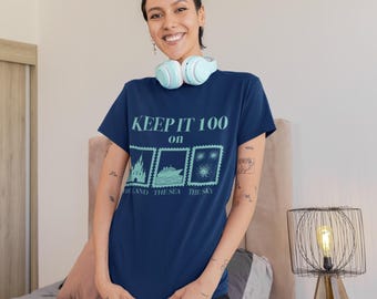 Puede incluir: Camiseta azul marino con el texto "KEEP IT 100" e ilustraciones de un castillo, un barco y fuegos artificiales. La camiseta también tiene las palabras "THE LAND THE SEA THE SKY". La persona lleva pantalones cortos negros y auriculares azul claro.