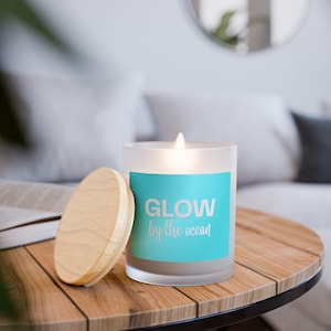 Puede incluir: Una vela encendida en un tarro de vidrio esmerilado con tapa de madera. El tarro tiene una etiqueta turquesa con las palabras "GLOW by the ocean" en blanco. La vela está sobre una mesa de madera.