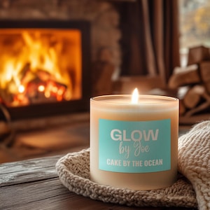 Puede incluir: Una vela encendida en un tarro de cristal transparente con una etiqueta azul claro que dice "GLOW by Joe CAKE BY THE OCEAN". La vela está sobre un posavasos de punto, con una manta acogedora cerca. Una chimenea con fuego ardiendo está en el fondo.