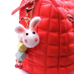 May include: A red quilted handbag with a white fluffy bunny keychain. The keychain has a gold ring and a multi-colored beaded charm.