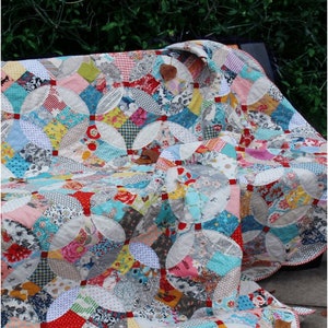May include: A colorful patchwork quilt with a white background and red, blue, and yellow accents. The quilt is folded and draped over a bench. The quilt is labeled "Magic Eye" and "Emma Mary Designs".