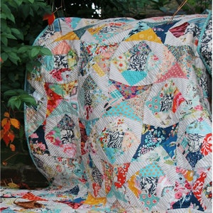 May include: A colorful quilt with a geometric pattern in shades of blue, pink, red, and yellow. The quilt is made of various fabrics with floral and animal prints. The quilt is folded and draped over a surface with green foliage in the background. The quilt is labeled "ARLINGTON SQUARE" and "EMMA MARY designs".