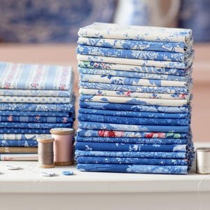 May include: Stacks of blue and white patterned fabric, featuring floral and striped designs. The fabrics are neatly folded and arranged on a white surface, with spools of thread and buttons nearby. Decorative blue and white china is visible in the background.