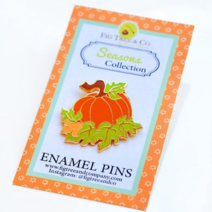 May include: Gold enamel pin featuring a pumpkin with green leaves and a brown stem. The pin is on a card with the text "Fig Tree & Co. Seasons Collection" and "Enamel Pins www.figtreeandcompany.com Instagram: @figtreeandco".