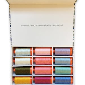 Op de afbeelding: Een doos met 12 grote spoelen 100% Aurifil katoenen garen. De spoelen zijn gerangschikt in een rasterpatroon en zijn in verschillende kleuren, waaronder blauw, groen, roze en geel. De doos is gelabeld "100% Aurifil Cotton &bull; 12 Large Spools &bull; 50wt &bull; 1422yds/spool".