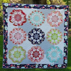 May include: A handmade quilt with nine floral medallion designs in shades of red, pink, blue, and green. The quilt has a navy blue floral border and a light gray background. The quilt is displayed outdoors on grass.