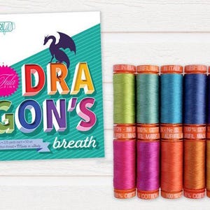 Op de afbeelding: Een verzameling kleurrijke garenspoelen naast een kaart met de tekst "DRAGON'S breath". De kaart toont een draak silhouet en de naam "Tula Pink". De garenspoelen zijn in verschillende tinten.