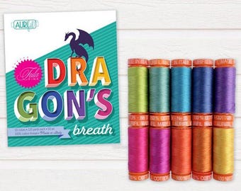Op de afbeelding: Een verzameling kleurrijke garenspoelen naast een kaart met de tekst "DRAGON'S breath". De kaart toont een draak silhouet en de naam "Tula Pink". De garenspoelen zijn in verschillende tinten.