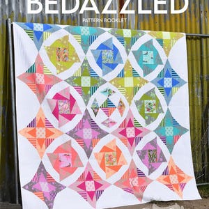 May include: A colorful quilt pattern booklet titled "Bedazzled" with a white background and a quilt design featuring a variety of bright colors and geometric shapes. The quilt is made up of squares, triangles, and circles in shades of pink, orange, blue, green, and yellow. The quilt is displayed against a rustic metal background.