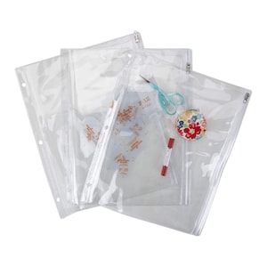 May include: Clear plastic zippered pouches with various craft supplies. The pouches are filled with items like scissors, a floral print container, and a red marker. Some pouches have printed text. The pouches are designed to hold craft supplies.