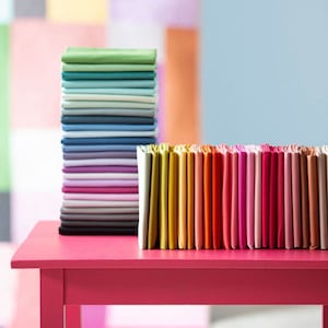 May include: A stack of colorful fabric swatches in various shades of green, blue, purple, pink, orange, and yellow. The swatches are arranged in a row on a pink table.