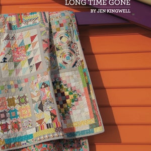Könnte beinhalten: Ein farbenfroher Patchwork-Decken mit verschiedenen Mustern und Stoffen, der an einer Holzwand mit violettem Rahmen hängt. Die Decke trägt den Titel "Long Time Gone" von Jen Kingwell.