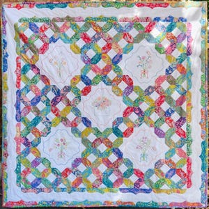 May include: A white quilt with a colorful patchwork design. The quilt features a repeating pattern of interlocking squares and rectangles. The squares and rectangles are made of different fabrics with floral prints in various colors, including pink, red, blue, green, and yellow. The quilt is edged with a white scalloped border.