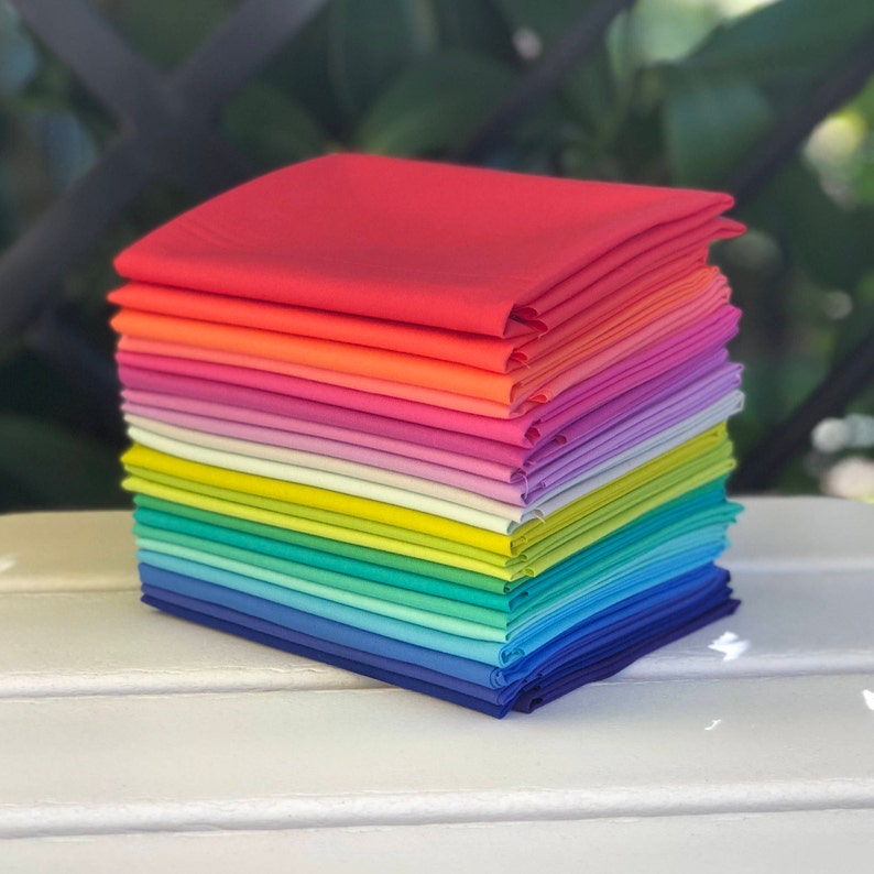 May include: A stack of 20 colorful fabric squares in a rainbow gradient, starting with red and ending with blue. The fabric is folded neatly and the stack is sitting on a white surface.