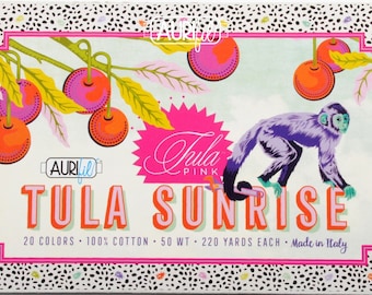 Op de afbeelding: Een doosje Aurifil Tula Pink garen. Het ontwerp toont een paarse aap, oranje en roze fruit en de woorden "Tula Sunrise". De doos heeft een witte achtergrond met een zwart-witte stippelrand en roze accenten.