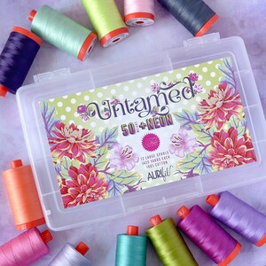 Op de afbeelding: Een transparante plastic opbergdoos met een kleurrijk bloemenontwerp en de tekst "Untamed 50+NEON" op de deksel. De doos bevat 12 grote spoelen Aurifil garen, elk met 1422 meter 100% katoen.
