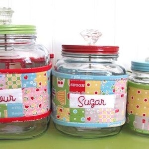 May include: Three glass jars with colorful patchwork fabric bands labeled "Flour", "Sugar", and "Tea". Each jar has a colored lid and a decorative glass knob. The jars are for kitchen storage.