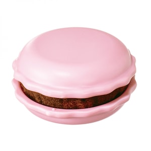 May include: A pink, round container shaped like a macaron, with a wavy edge. The top and bottom are a solid, light pink, and the filling is dark brown. The design mimics a French macaron pastry.