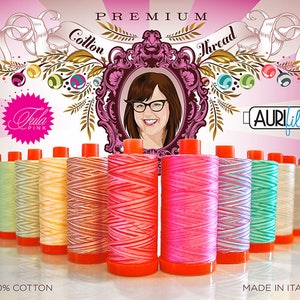 May include: A collection of 10 spools of Aurifil thread in various colors and patterns. The thread is 100% cotton and made in Italy. The spools are arranged in a row on a white surface. The text "PREMIUM Cotton Thread" is visible at the top of the image. The text "Tula Pink" is visible in a pink starburst shape. The text "Aurifil" is visible in a white rectangle with a black border.