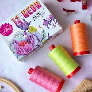 May include: A box of 12 weight Aurifil thread in neon colors with the Tula Pink logo. The box is open and three spools of thread are visible: pink, green, and orange.