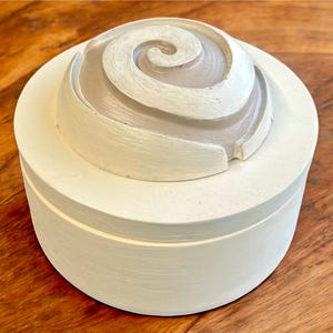 May include: A white, round decorative box with a spiral design on the lid. The lid's spiral is a lighter shade of white, creating a subtle contrast. The box sits on a wooden surface.