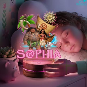 May include: A night light featuring characters from the animated movie Moana, including Maui, Moana, and Pua. The name "SOPHIA" is displayed in pink letters. The light sits on a wooden base and emits a soft glow.