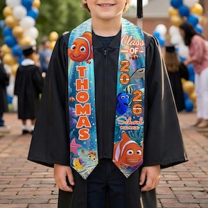 May include: A person in a black graduation gown and cap, wearing a custom sash. The sash is light blue with orange text that reads "THOMAS" and "Class of 2026." The sash features cartoon fish illustrations. The background shows a graduation celebration.
