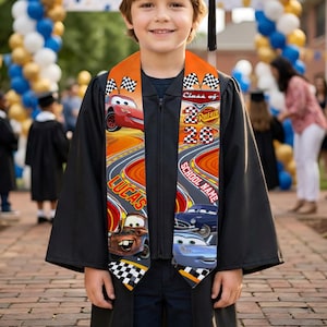 May include: A young person wearing a black graduation gown and cap with a custom orange and black graduation sash. The sash features the text "Class of 2023" and "Cars" characters, with the name "Lucas" printed on it.